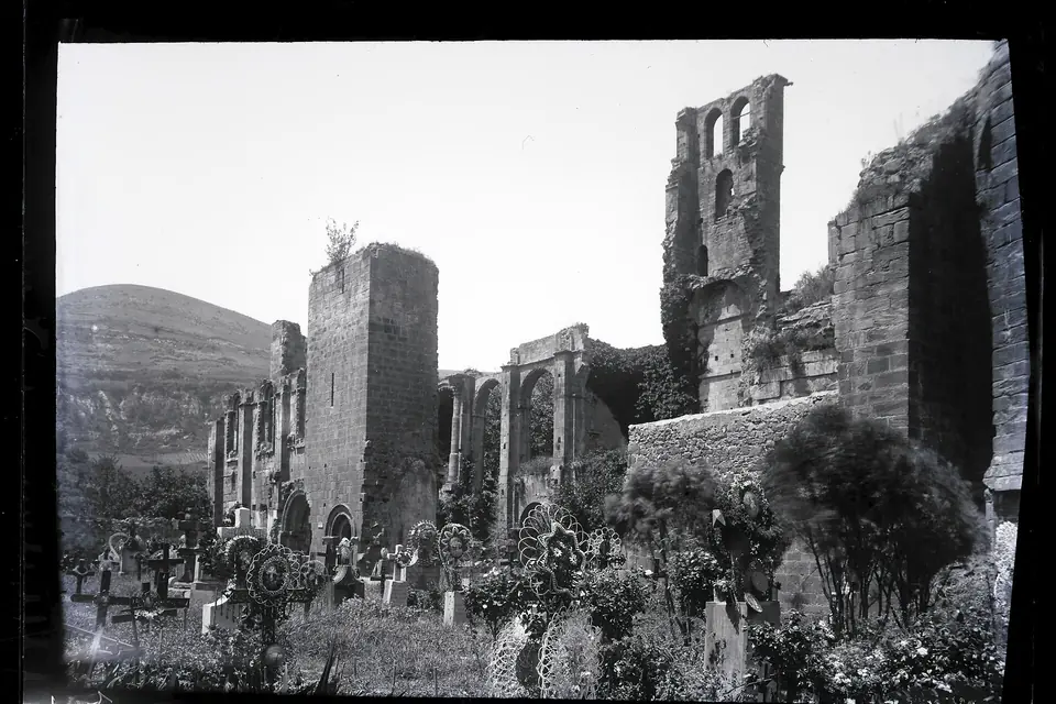 Photographie ancienne en noir et blanc de ruines d'église envahies de végétation, avec des pans de murs et une tour, au pied d'une colline