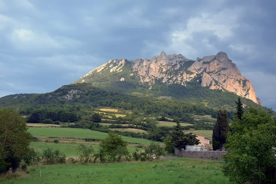 Une montagne calcaire aux falaises claires dominant une vallée verte et boisée, sous un ciel d'été avec quelques nuages