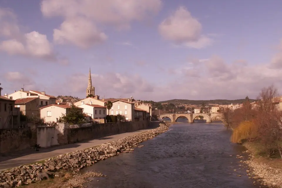 La rivière Aude traversant une petite ville aux maisons claires, avec un clocher pointu et un pont de pierre à arches au fond, sous un ciel de fin de journée