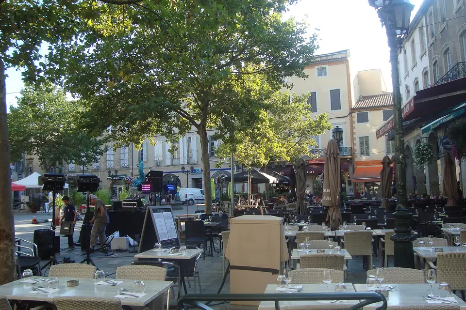 Une place ombragée de platanes, bordée de façades claires et de terrasses de café, sous le soleil