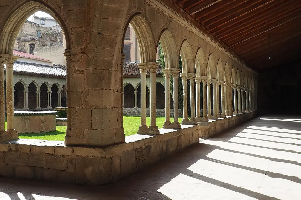 Galerie de cloître aux arcs brisés sur colonnettes jumelées, sous une charpente de bois, avec un jardin ensoleillé au centre